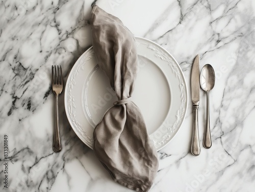 A white plate with a knotted beige napkin, flanked by a fork, knife, and spoon, on a marble surface