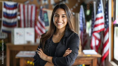 Smiling young hispanic woman with arms crossed standing in a room decorated with american flags and a voting booth indicating an election setting in the united states isolated on white background, rea