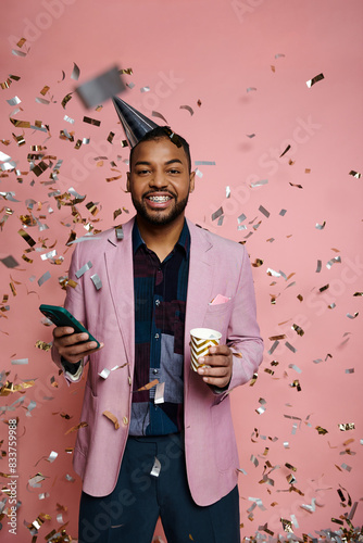 Young African American man in party hat and braces holds cup and cell phone against pink background.