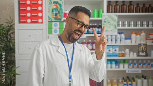 A cheerful hispanic male pharmacist winking and gesturing peace in a well-stocked modern pharmacy.