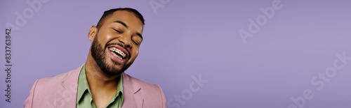 Young African American man with braces laughing joyfully in a stylish pink jacket against a vibrant purple backdrop.