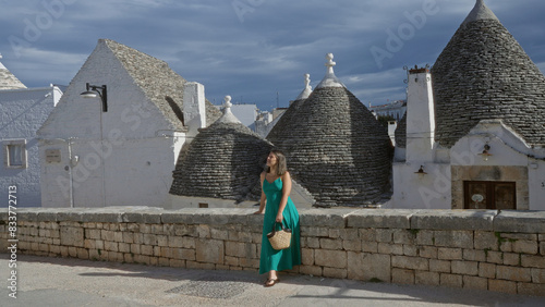 Beautiful young hispanic woman in a green dress stands on a cobblestone street in the old town of alberobello, italy, admiring the unique trulli houses under a dramatic sky.