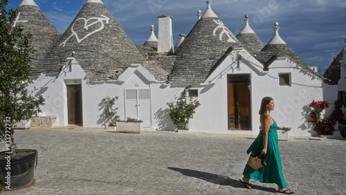 Young hispanic woman in a green dress walking past traditional trulli houses in the old town of alberobello, puglia, italy on a sunny day.