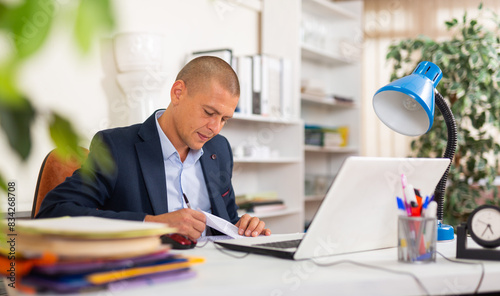 Positive businessman working with laptop and papers at office desk