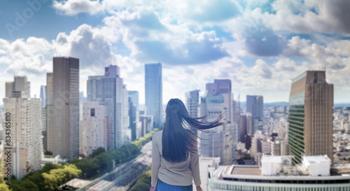 日本の街・都市・朝日を受けたオフィスビル群を前に佇み風を受けて髪をなびかせる会社員の女性の出勤 ビジネス・はじまり・決意・目標・挑戦のイメージ