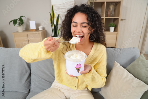 Beautiful young African-American woman eating ice cream and sitting on sofa at home