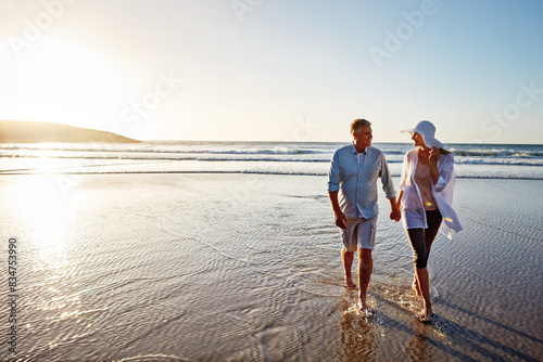 Mature couple, holding hands and walking with beach sunset for love, embrace or bonding together in nature. Man, woman or lovers enjoying stroll on water for outdoor holiday or summer by ocean coast