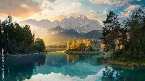 Painterly lake scenery in Germany with mountains reflected in the teal water and a beautiful ray of morning sunlight illuminating some trees 