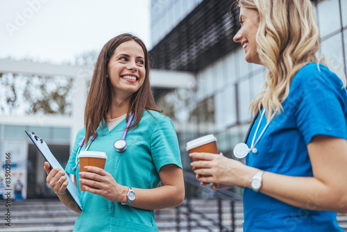 Two smiling female nurses in blue scrubs relax with coffee cups during a break outside a medical facility, enjoying a moment of camaraderie.