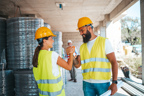 A smiling male and female construction worker shake hands on a busy construction site, attired in safety vests and hard hats, reflecting teamwork and collaboration in a professional setting.