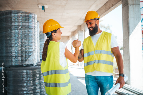 A male and female worker in safety vests and helmets engage in a firm handshake, exemplifying teamwork on a construction site.