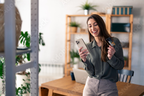 Happy young woman holding credit card using cell phone mobile app paying online making purchase in e-commerce digital store on smartphone at home. Virtual shopping, secure mobile banking concept.