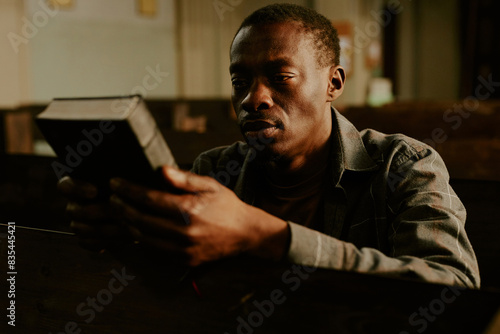 Young African American man sitting on bench in Catholic church holding Bible book in hands, medium closeup shot