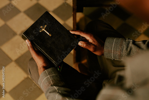 High angle view of hands of unrecognizable Black man sitting on pew in Catholic church holding prayer book