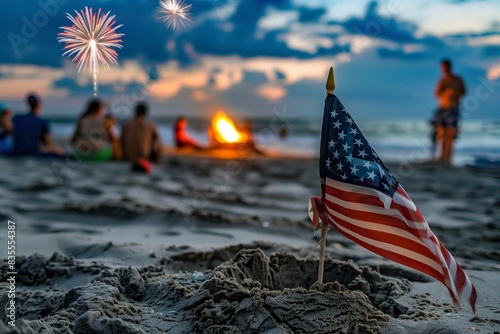  A small American flag is planted in the sand on a beach, with a blurred background featuring a bonfire and people gathered in USA during independence day. 