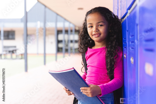 In school, outdoors, young biracial girl holding a book stands by lockers, copy space