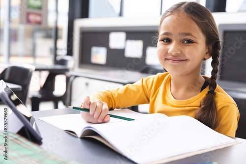 In school, in a classroom, a young biracial girl with light brown skin is writing in a notebook