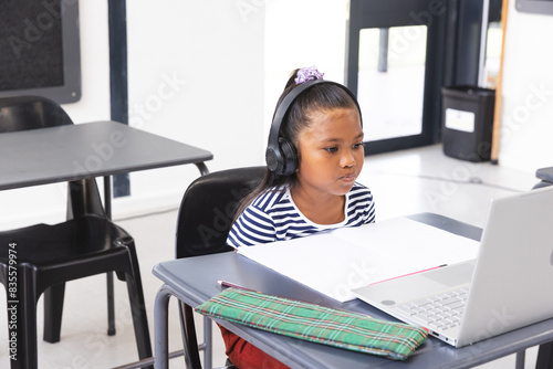 In school, young biracial female student wearing headphones focusing on a laptop in the classroom