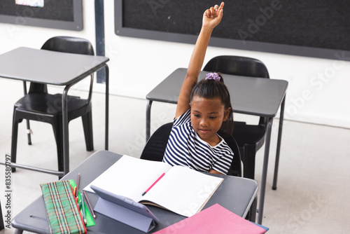 In school, young biracial girl with brown skin and dark hair is raising her hand in the classroom