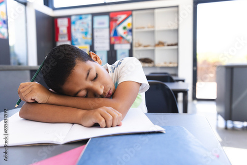 Exhausted biracial boy sleeps on desk in bright classroom.