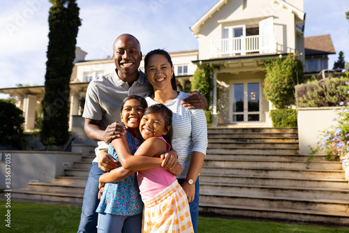 Outdoors, diverse family of four smiling and embracing in front of their home