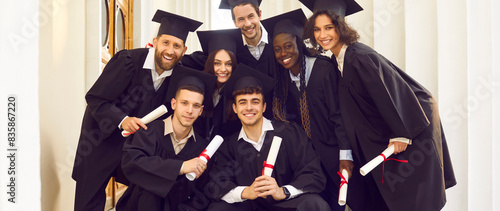Portrait of a smiling happy multiracial international graduates students standing in a university graduate gown and holding diploma outdoor. Education and graduation concept. Banner.