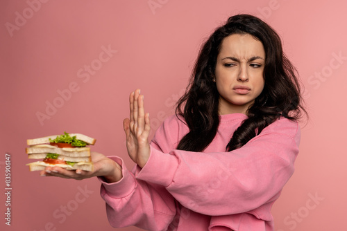 Displeased young dark-haired female showing her food aversion during the studio photo shoot on the pink background. Self-control and nutrition concept