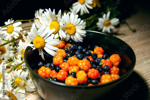 Cloudberry & blueberry (bilberry, whortleberry, huckleberry, blaeberry) in bowl with white flowers daisies. Cloudberry berry mix. Wild chamaemorus berries organic on wooden background in summer day