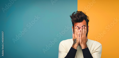 A man with tattoos on his arm. The background is a mix of orange and blue colors. Man's facial expression. a handsome italian man feeling disappointed and covers his face with both his hands
