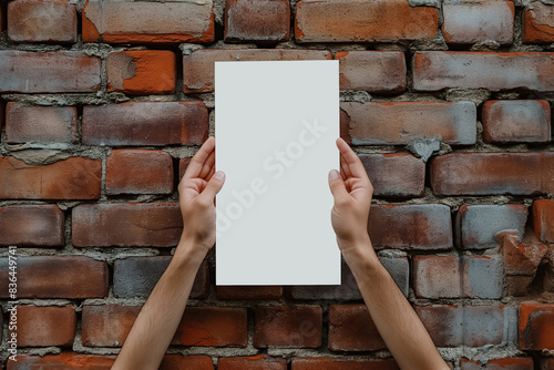 Hands holding a white blank paper vertically against a red brick wall background; mockup