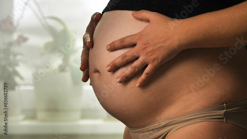 Third trimester of pregnancy. A pregnant woman gently cuddles her developed belly, wearing beige underwear and an elevated black top. The background is a blur of indoor plants on a windowsill.