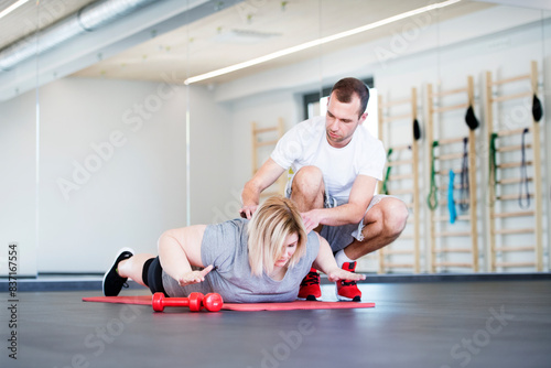 Overweight woman exercising on gym mat in gym. Personal trainer couching her and helping her.