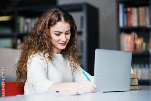 Young female student in library, focusing on final project, presentation, working on laptop. University student preparing for final exam.