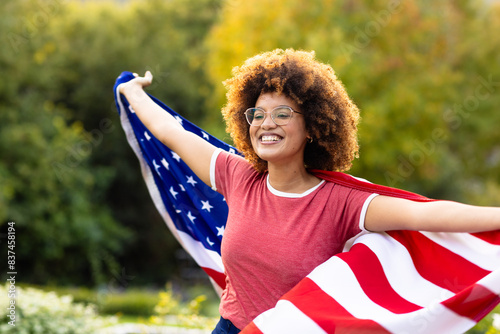 Happy biracial woman covered with flag of usa with arms wide in garden