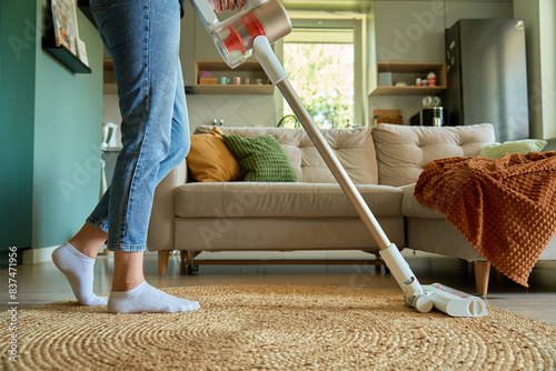 Woman is vacuuming floor of modern apartment living room, using portable cordless vacuum cleaner. Daily routine and household chores