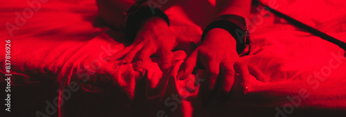 hands in BDSM handcuffs of a submissive female slave on a sheet on a bed with a red light. Wide horizontal banner header