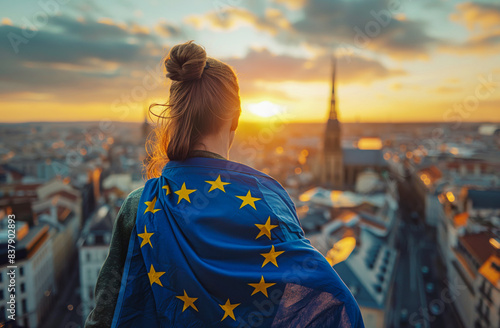 Rear view of female person with flag of European Union on her shoulders looking at sundown above city.