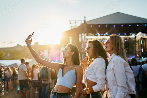 Girls in summer attire enjoy sunset at coastal music festival, stage lights glow in background. Youth share moments, dance to live performances, sandy beach venue vibrates with fest energy.