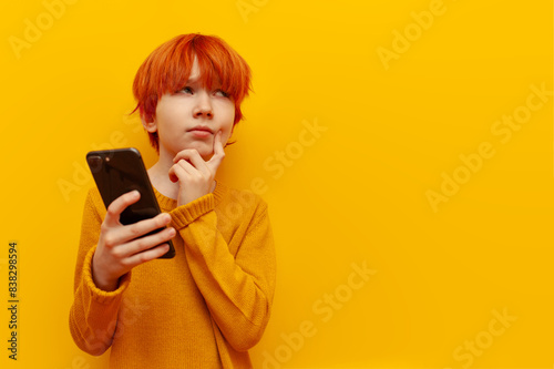 pensive red-haired child boy using smartphone and planning on yellow isolated background, puzzled teenager winner with orange hairstyle thinks and imagines and looks away