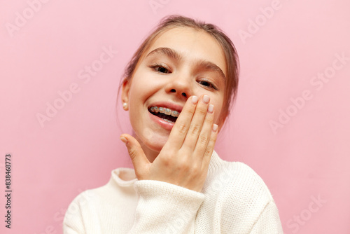 cheerful teenage girl with braces mocks and covers smile with hand over pink isolated background, embarrassed child feels shy and laughs