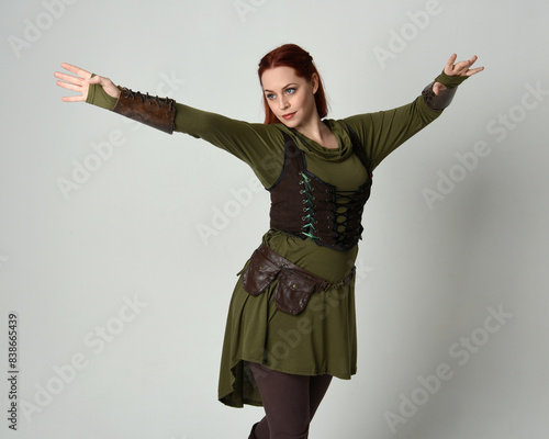 Close up portrait of beautiful red haired female model, wearing green and brown medieval fantasy costume with tunic and armour. Standing with gestural hand poses, isolated on white studio background