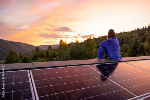 Rooftop with solar panels on house in mountains, woman sitting alone enjoying sunset. Energy independence, sustainability, self sufficient, and escapism to nature concept