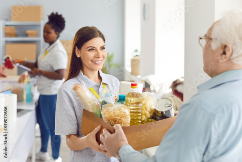 Positive volunteer woman giving a food donation box to a pensioner at a social assistance center or organization. Charity work providing humanitarian help and social support to those in need.