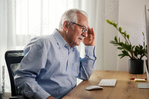 A senior man sitting at home office with computer and having headache.
