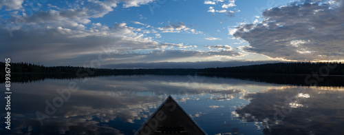 A panoramic view from a canoe at a large calm northern lake in the early morning. The front of the boat is in the center of the frame. The calm water reflects the white to gray clouds in a blue sky.