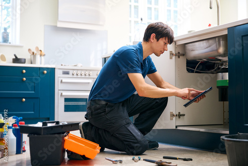 Male Plumber With Digital Tablet Fixing Waste Disposal Unit In Domestic Kitchen Sink