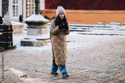 A young Ukrainian girl in a down jacket, an Angora hat and gloves walks along a snowy winter street and looks at a smartphone. Lviv, Ukraine.