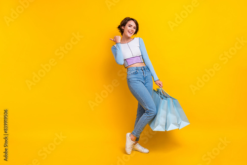 Full body photo of charming cheerful lady hold mall bags indicate finger empty space isolated on yellow color background