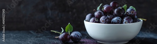 Deep purple sloe berries in a white ceramic bowl, with one fruit opened to show the juicy, dark interior, set against a dark background