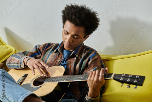 A young African American man sitting on a couch, playing an acoustic guitar while talking on the phone.